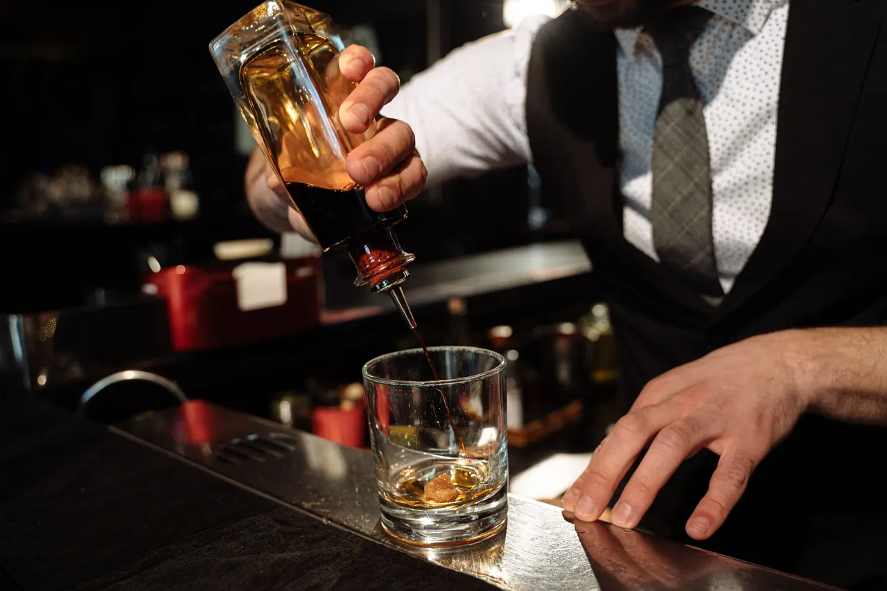 A bartender pouring an amber spirit into a glass over ice at the bar